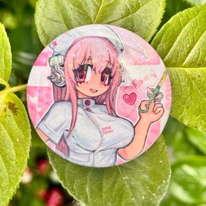 以下が含まれることがあります： ピンク色の髪をしたアニメ風のキャラクターが、白いナース服とナースキャップを着用している円形の缶バッジ。キャラクターは注射器を持っており、背景はハートが描かれたピンク色。ナース服には「SUPER SONICO」という文字がプリントされている。