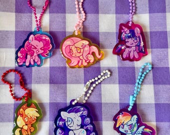 May include: A collection of six colorful acrylic keychains featuring cartoon pony characters. Each keychain has a different pony design with vibrant colors, attached to a beaded chain. The keychains are displayed on a purple and white checkered background.