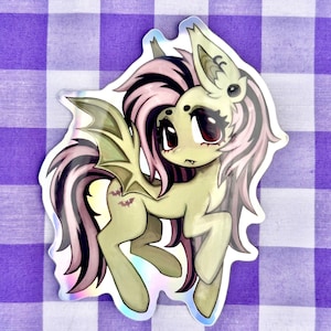 May include: A die-cut sticker featuring a pale green pony with large dark eyes, a single fang, and bat-like wings. The pony has a flowing mane and tail with pink and black streaks, and two small purple bat silhouettes on its flank, all with a holographic finish.