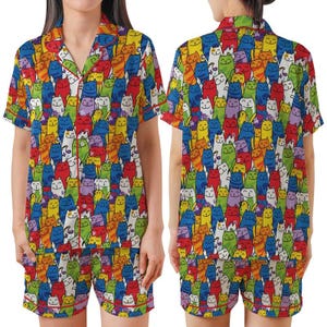 May include: A colorful pajama set featuring a vibrant pattern of cartoon cats in various colors. The set includes a short-sleeved button-down shirt and matching shorts, both trimmed with red piping. The cat design covers the entire set.