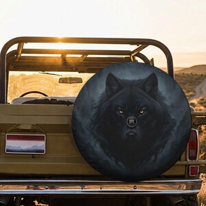 Op de afbeelding: Een ronde reservewielhoes met een gedetailleerde afbeelding van een zwarte wolf met gloeiende amberkleurige ogen. De wolf is gecentreerd tegen een donkere, mistige bosachtergrond. De hoes is gemonteerd op de achterkant van een olijfgroen terreinvoertuig.