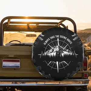 May include: A black circular spare tire cover featuring a white graphic of a mountain range, a forest, and a compass rose. The text "ALWAYS TAKE THE SCENIC ROUTE" is printed in a white arc above the mountain illustration on the cover.