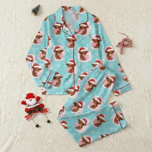Puede incluir: Conjunto de pijama azul claro con un estampado repetido de vacas de las Tierras Altas de dibujos animados con gorros de Papá Noel y soplando burbujas de chicle. El conjunto incluye una camisa con botones y pantalones a juego, ribeteados con ribetes rojos. Ropa de dormir navideña festiva.