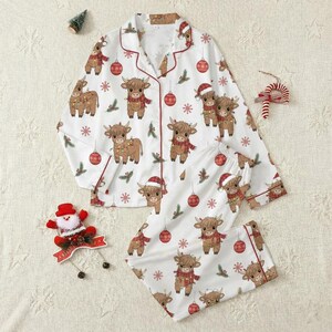 Peut inclure: Ensemble de pyjama blanc sur le thème de Noël, avec un motif répété de vaches des Highlands de dessin animé portant des écharpes et des bonnets de Père Noël, avec des boules de Noël et des flocons de neige. L'ensemble comprend une chemise boutonnée et un pantalon.