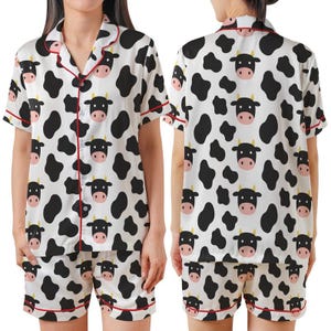 Puede incluir: Conjunto de pijama blanco con estampado de vaca negra y ribete rojo. La parte superior de manga corta tiene una parte delantera abotonada y un cuello con muescas. Los pantalones cortos a juego tienen cintura con cordón. El estampado de vaca presenta manchas negras y caras de vaca.