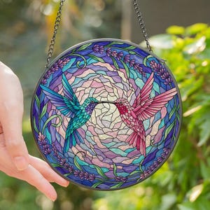 Könnte beinhalten: Ein rundes Buntglasfenster mit zwei Kolibris. Einer ist türkis, der andere rosa, mit sich berührenden Schnäbeln. Der Hintergrund ist ein Mosaik aus lila, blauem und rosa Glas, umgeben von Lavendelblüten. Eine Metallkette ist zum Aufhängen befestigt.
