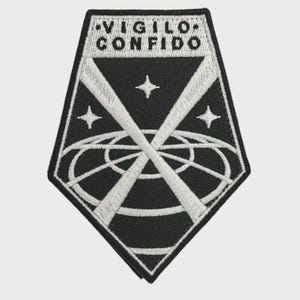 May include: Black and white embroidered patch with the text "VIGILO CONFIDO". The patch features a geometric design with intersecting lines, stars, and a circular element, creating a striking visual.