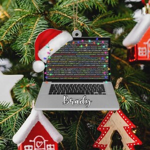 May include: A Christmas tree ornament shaped like a laptop with the name "Brady" in cursive. The laptop screen displays code. A Santa hat and string lights are attached to the ornament. Other Christmas ornaments are visible.