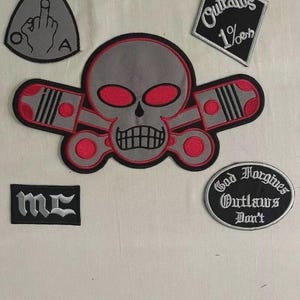 May include: A collection of embroidered patches with biker-themed designs. Includes a skull with pistons, a diamond-shaped patch with "Outlaws 1%er", and an oval patch with "God Forgives Outlaws Don't".