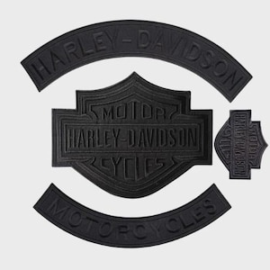 May include: Black Harley-Davidson patches in various shapes. The set includes a large shield, two arched patches with the brand name, and a smaller shield-shaped patch. The patches are made of a textured material.
