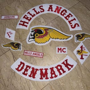 May include: A collection of embroidered patches featuring the words "HELLS ANGELS" and "DENMARK" in red lettering on white backgrounds. The patches include a skull with wings and the letters "MC" and "1%" symbols.