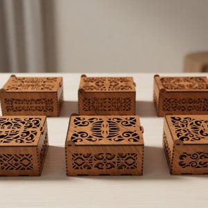 Può includere: Sei piccole scatole quadrate in legno con intricati disegni tagliati al laser sui coperchi e sui lati. Le scatole sono di un caldo tono legno naturale e sono disposte su una superficie chiara. Scatole decorative per piccoli oggetti.