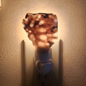 Puede incluir: Una luz de noche decorativa con un diseño natural y orgánico. La luz presenta una pantalla de color marrón y beige, similar a una piedra, que emite un brillo cálido cuando se enchufa a una toma de corriente. La luz de noche está hecha de un material translúcido.