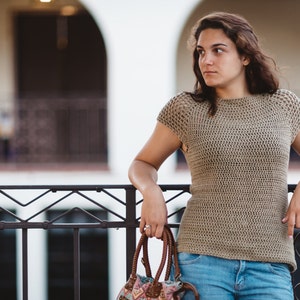 May include: A woman wearing a beige crochet top and blue jeans stands in front of a railing. She is holding a brown and floral patterned handbag.