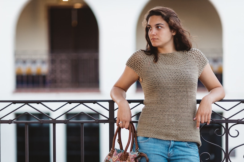 May include: A woman wearing a beige crochet top and blue jeans stands in front of a railing. She is holding a brown and floral patterned handbag.