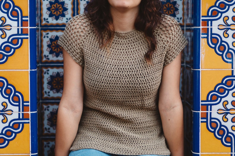 May include: A woman wearing a brown crochet top with a loose fit and short sleeves. The top has a textured, open-weave design. The woman is standing in front of a wall with colorful tile patterns.