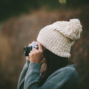 Könnte beinhalten: Eine Person trägt eine weiße Strickmütze mit Bommel und einen grauen Pullover und hält eine Vintage-Kamera in der Hand, um ein Foto zu machen.