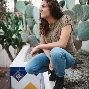 May include: A woman wearing a brown crocheted top and blue jeans sits on a white ledge. The ledge is decorated with blue and yellow tiles. The woman is looking to the right of the frame.