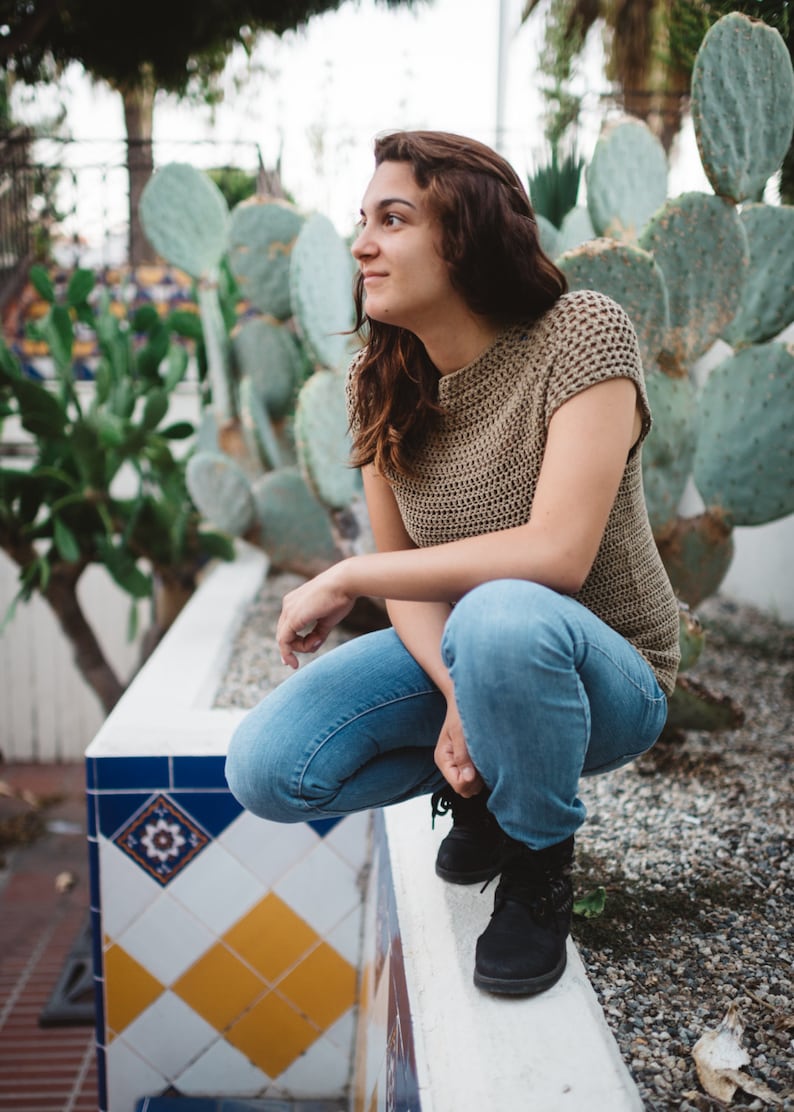 May include: A woman wearing a brown crocheted top and blue jeans sits on a white ledge. The ledge is decorated with blue and yellow tiles. The woman is looking to the right of the frame.