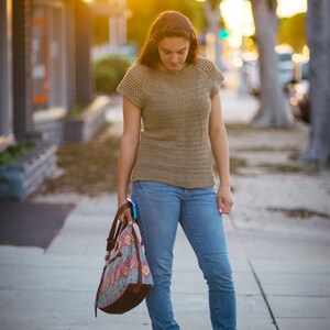 May include: A woman wearing a brown crochet top and blue jeans is walking down a sidewalk. She is carrying a colorful patterned tote bag.
