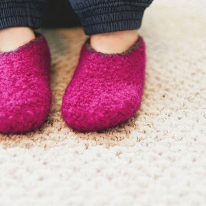 May include: A pair of fuzzy pink slippers with a dark grey trim. The slippers are on a light beige carpet.