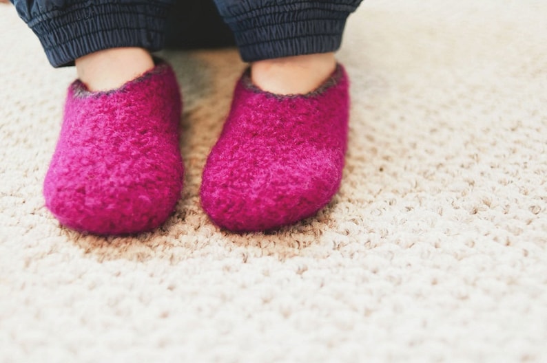 May include: A pair of fuzzy pink slippers with a dark grey trim. The slippers are on a light beige carpet.