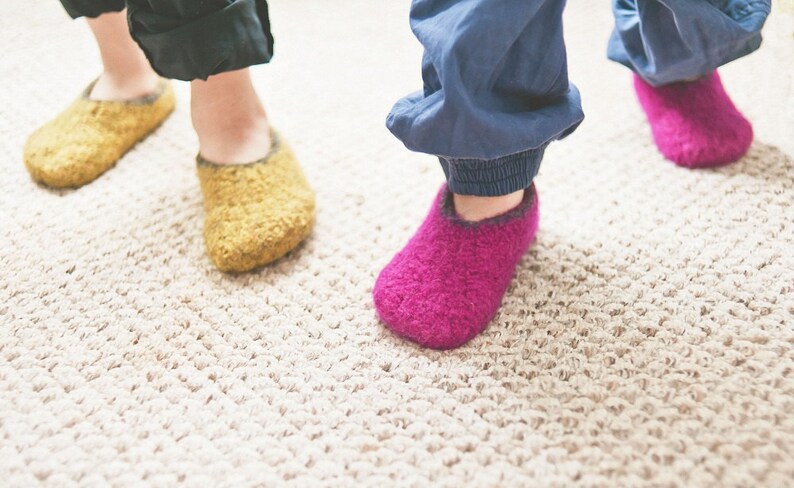 May include: Two pairs of fuzzy slippers, one pair is yellow and the other is pink. The slippers are on a light beige carpet.