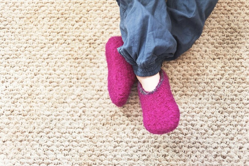 May include: A pair of fuzzy pink slippers with a dark gray trim. The slippers are worn by a person sitting on a beige carpet.