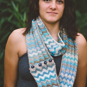 May include: A woman wearing a blue, white, and brown crocheted infinity scarf with button accents. The scarf has a chevron pattern.