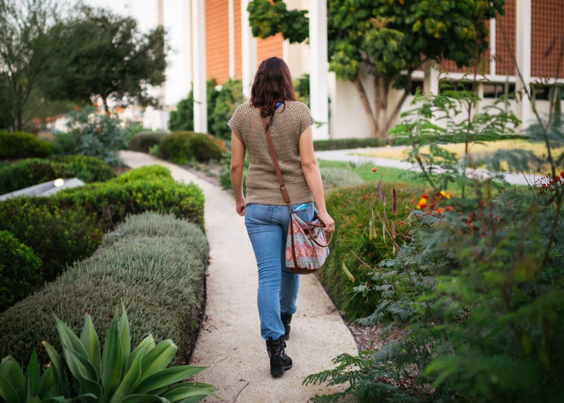 May include: A woman walks down a paved path in a garden setting. She is wearing a brown crocheted top, blue jeans, and a brown leather shoulder bag with a colorful patterned design. The path is lined with lush green bushes and plants.