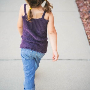 May include: A young person wearing a purple knitted tank top and blue jeans walks down a pavement. They have their hair pulled back in two pigtails with yellow hair ties.