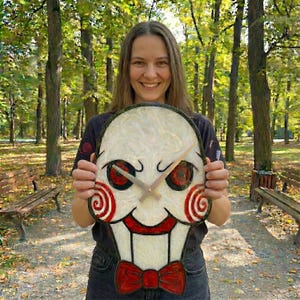 May include: A decorative wall clock featuring a stylized face with red and white spiral cheeks, red eyes, and a red bow tie. The clock has beige clock hands and a white background. The clock is held in front of a person in a park setting.