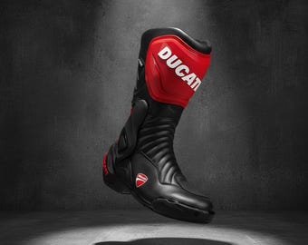 May include: Black and red motorcycle boot with the word "DUCATI" in white lettering. The boot has a high-top design with a padded upper and a textured, ribbed lower section. The Ducati logo is visible on the side of the boot.
