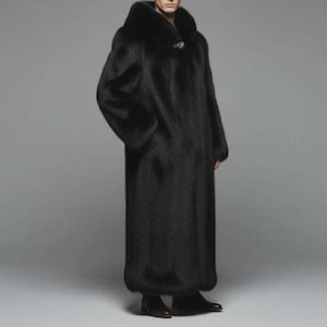 May include: A full-length black faux fur coat with a large collar and a decorative clasp. The coat has long sleeves and a tailored fit. The coat is worn with black shoes. The background is a solid gray.