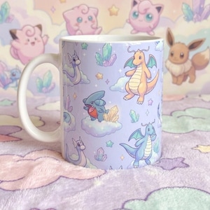 Könnte beinhalten: Ein weißer Keramikbecher mit einem sich wiederholenden Muster auf hellviolettem Hintergrund. Das Design zeigt verschiedene cartoonartige drachenähnliche Kreaturen, kleine Wolken und bunte Kristalle. Der Henkel ist weiß und links angebracht.