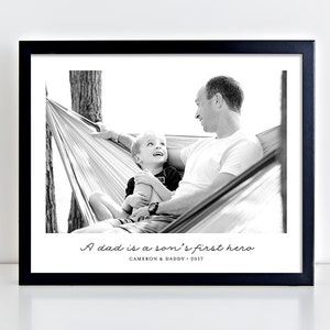 May include: Black and white photograph of a father and son in a hammock. The father is smiling and looking at his son, who is also smiling. The caption reads "A dad is a son's first hero. Cameron & Daddy - 2017."
