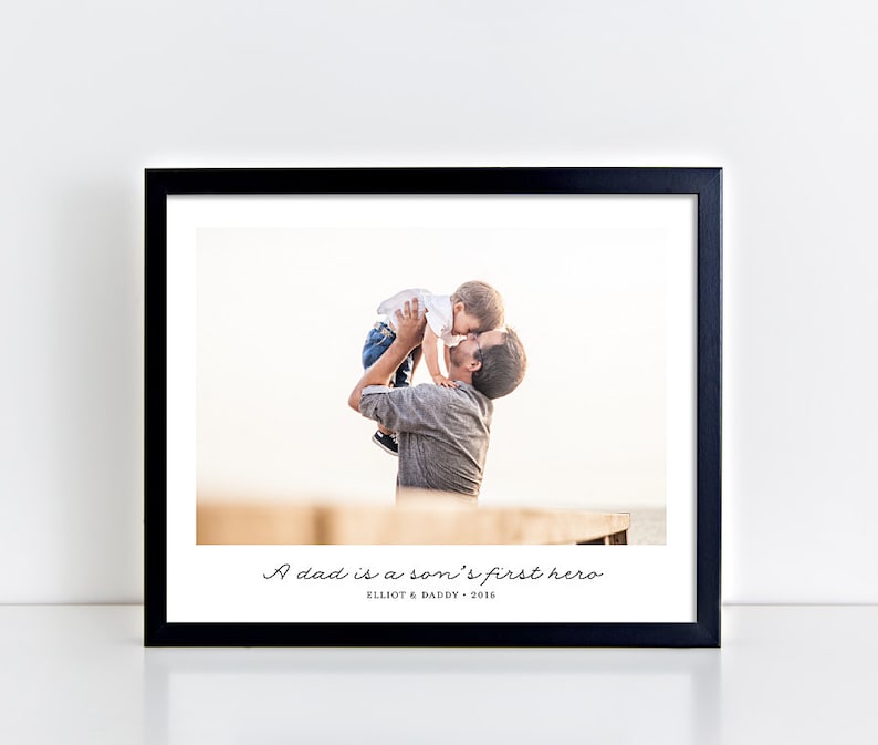 May include: A black framed photograph featuring a father and son. The father is holding the son aloft and kissing him on the cheek. The photograph includes the text "A dad is a son's first hero" and "Elliot & Daddy - 2016" printed beneath the image.