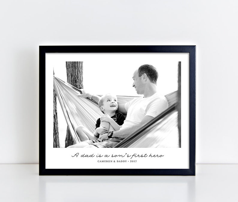 May include: Black and white photograph of a father and son in a hammock. The father is smiling and looking at his son, who is also smiling. The caption reads "A dad is a son's first hero. Cameron & Daddy - 2017."