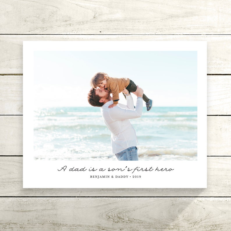 May include: A father and son on a beach. The father is holding the son in the air and they are both smiling. The text "A dad is a son's first hero" is written below the image. The text "Benjamin & Daddy - 2019" is written below the first text.