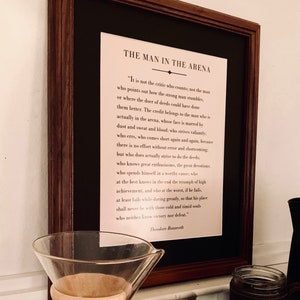 May include: A framed print with a black background and white text. The text is a quote by Theodore Roosevelt about "The Man in the Arena".