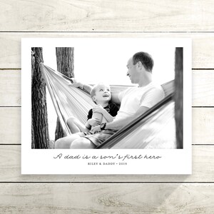May include: A black and white photograph of a father and son in a hammock. The father is smiling and looking at his son, who is looking up at him. The text "A dad is a son's first hero" is written below the photo, along with the names "Riley & Daddy" and the year "2019".