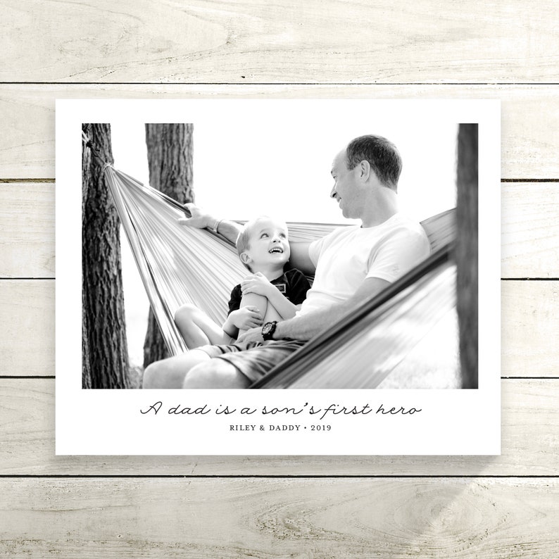 May include: A black and white photograph of a father and son in a hammock. The father is smiling and looking at his son, who is looking up at him. The text "A dad is a son's first hero" is written below the photo, along with the names "Riley & Daddy" and the year "2019".