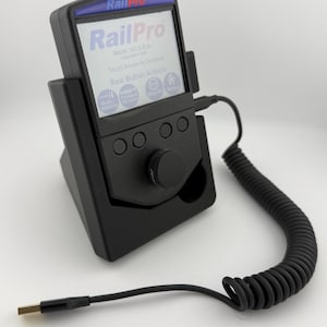 Peut inclure: Appareil RailPro noir avec un écran affichant le logo RailPro et du texte. L'appareil est dans un socle noir avec un cordon spiralé et un connecteur USB. L'écran affiche "Touch Screen to Continue" et "Real Button Actions".