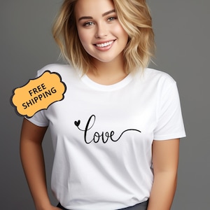 Può includere: T-shirt bianca con un design nero della parola "Love" in corsivo e un piccolo cuore. La maglietta è a girocollo e sembra essere fatta di un materiale morbido. La persona che indossa la maglietta ha i capelli biondi.