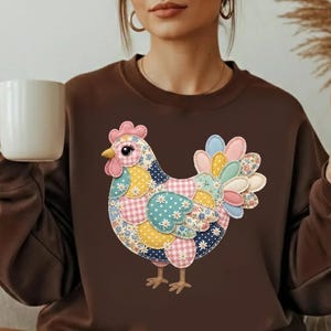 Könnte beinhalten: Ein braunes Sweatshirt mit einem bunten Patchwork-Hühnerdesign. Das Huhn hat einen rosa Kamm, einen gelben Schnabel und einen mehrfarbigen Schwanz. Das Sweatshirt ist ein bequemer, lässiger Stil.