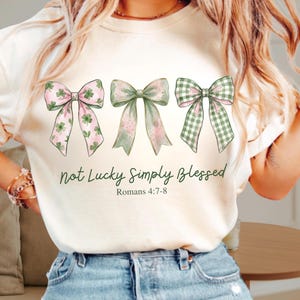 Puede incluir: Camiseta color crema con tres lazos decorativos en tonos verdes y rosas. Los lazos tienen diseños de tréboles y cuadros. El texto "Not Lucky Simply Blessed" y "Romans 4:7-8" está impreso debajo de los lazos.
