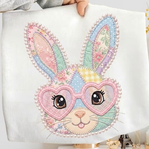 Peut inclure: Un tissu blanc avec un visage de lapin patchwork portant des lunettes roses en forme de cœur. Le lapin a des yeux bruns, un nez rose et une bordure de perles. Les oreilles sont décorées de motifs floraux et géométriques.