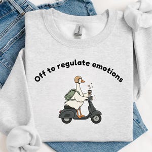 May include: Light gray sweatshirt with the text "Off to regulate emotions." A cartoon goose wearing a helmet and backpack rides a black scooter while holding a coffee cup. The sweatshirt is paired with blue jeans.