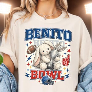 May include: A cream-coloured t-shirt with a cartoon bunny holding a trophy, and the text "BENITO BOWL" in blue and red. The design includes a football, lightning bolts, stars, a foam finger, and a football helmet.