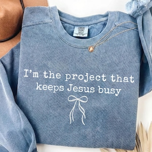 Puede incluir: Sudadera azul de cuello redondo con el texto "I'm the project that keeps Jesus busy" en blanco. Un collar dorado con un colgante está sobre la sudadera. Un gráfico de lazo blanco está debajo del texto.